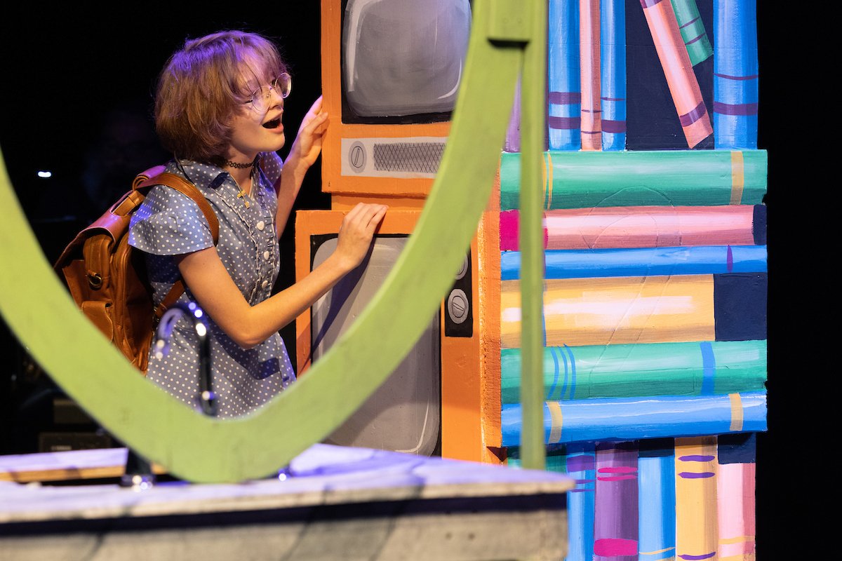 Young student peering around stylized bookcase and singing on stage in Matilda